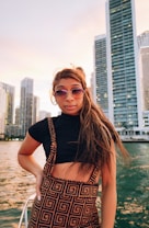 A person stands confidently in front of a body of water with modern skyscrapers in the background. They are wearing stylish sunglasses and a patterned outfit, with long hair blowing gently in the wind. The sky has a warm, soft glow, suggesting a sunset.