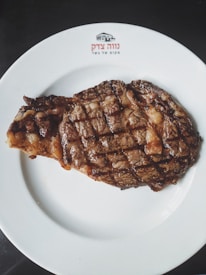 A grilled steak with a charred exterior is placed on a white plate. The plate has a logo and text written on it, possibly in Hebrew. The steak is cooked with visible grill marks and appears juicy and well-seasoned.