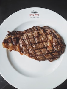A grilled steak with a charred exterior is placed on a white plate. The plate has a logo and text written on it, possibly in Hebrew. The steak is cooked with visible grill marks and appears juicy and well-seasoned.