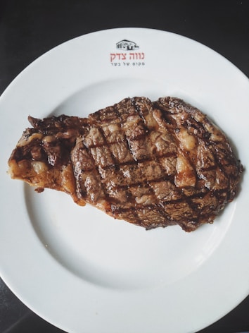 A grilled steak with a charred exterior is placed on a white plate. The plate has a logo and text written on it, possibly in Hebrew. The steak is cooked with visible grill marks and appears juicy and well-seasoned.
