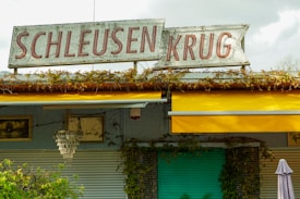 The exterior of a building features a vintage sign with the words 'Schleusen Krug' in large red letters. A yellow awning provides shade below the sign, and climbing plants are growing on the structure. The wall is adorned with framed decorations, and a light fixture resembling a chandelier is also visible.