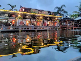 A vibrant lakeside restaurant with warm lighting, featuring a thatched roof structure and palm trees in the background. The ambient light reflects beautifully on the water's surface, creating a serene and inviting atmosphere. Groups of people are seen enjoying the evening under the shelter, adding to the lively yet relaxed setting.