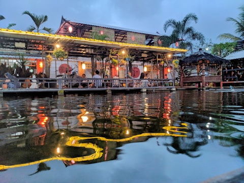 A vibrant lakeside restaurant with warm lighting, featuring a thatched roof structure and palm trees in the background. The ambient light reflects beautifully on the water's surface, creating a serene and inviting atmosphere. Groups of people are seen enjoying the evening under the shelter, adding to the lively yet relaxed setting.