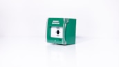 A green emergency door release box with a white front panel and a button labeled 'Press Here'. The box appears to be made of plastic and is set against a plain white background.