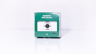 A green emergency door release box with a clear cover and text indicating 'Emergency Door Release' and 'Press Here' with an arrow pointing towards a central black dot.