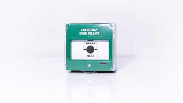 A green emergency door release box with a clear cover and text indicating 'Emergency Door Release' and 'Press Here' with an arrow pointing towards a central black dot.