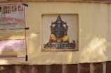 A painting of a deity with an elephant head and a yellow-orange robe is set within a recessed niche on a beige wall. Adjacent to it is a colorful poster with text in a script that appears to be Hindi, taped to the wall.
