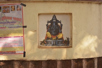 A painting of a deity with an elephant head and a yellow-orange robe is set within a recessed niche on a beige wall. Adjacent to it is a colorful poster with text in a script that appears to be Hindi, taped to the wall.