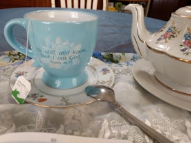 A blue ceramic mug with an inspirational quote sits on a decorative plate alongside a floral-patterned teapot. A tea bag with a green and white tag hangs from the mug, and a metal spoon rests on the saucer. The setting includes a tablecloth with intricate lace designs and a hint of a book at the bottom.