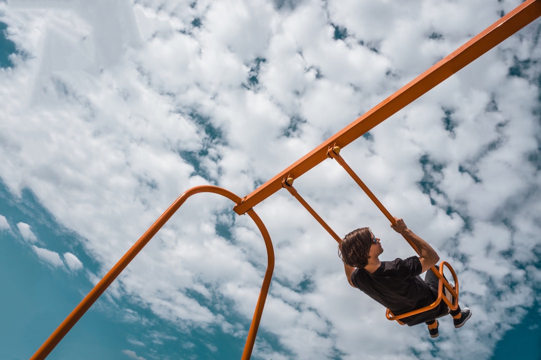 man in black jacket and blue denim jeans sitting on orange metal bar under blue and, Swinging in the sky