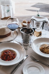 A dining table is elegantly set with two plates of pasta, one topped with tomato sauce and the other with olive oil and vegetables. A glass of red wine and a glass of white wine accompany the meal, alongside a partially visible beer bottle resting in an ice bucket. A straw hat is placed nearby, adding a casual touch to the sophisticated setting. In the background, a bread basket and a bottle of sparkling water are seen.