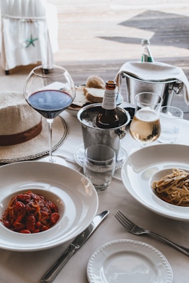 A dining table is elegantly set with two plates of pasta, one topped with tomato sauce and the other with olive oil and vegetables. A glass of red wine and a glass of white wine accompany the meal, alongside a partially visible beer bottle resting in an ice bucket. A straw hat is placed nearby, adding a casual touch to the sophisticated setting. In the background, a bread basket and a bottle of sparkling water are seen.