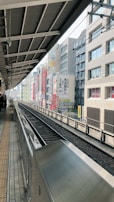 A railway platform with tracks on the right side, lined by urban buildings partially visible in the background. Overhead structures of the station are prominent, along with signage in a non-Latin script. The setting appears to be in a city, with no people visible on the tracks.