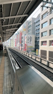 A railway platform with tracks on the right side, lined by urban buildings partially visible in the background. Overhead structures of the station are prominent, along with signage in a non-Latin script. The setting appears to be in a city, with no people visible on the tracks.