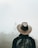 A person wearing a cowboy hat and a backpack faces away, looking at a distant, fog-shrouded temple. The scene is misty, creating an ethereal atmosphere.