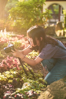 A person is focused on taking a photograph of vibrant plants with a camera mounted on a tripod. The setting appears to be a garden with lush greenery and sunlight filtering through the foliage, creating a serene and warm atmosphere.