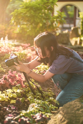 A person is focused on taking a photograph of vibrant plants with a camera mounted on a tripod. The setting appears to be a garden with lush greenery and sunlight filtering through the foliage, creating a serene and warm atmosphere.