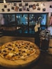 A thin-crust pizza topped with various ingredients including sausage, peppers, and cheese is placed on a wooden board. The setting is a dimly lit cafe, featuring hanging lightbulbs and an arrangement of photographs clipped to a string on the wall. A reflective French press and a dark bottle are situated next to the pizza.