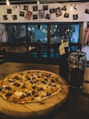 A thin-crust pizza topped with various ingredients including sausage, peppers, and cheese is placed on a wooden board. The setting is a dimly lit cafe, featuring hanging lightbulbs and an arrangement of photographs clipped to a string on the wall. A reflective French press and a dark bottle are situated next to the pizza.