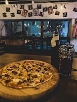 A thin-crust pizza topped with various ingredients including sausage, peppers, and cheese is placed on a wooden board. The setting is a dimly lit cafe, featuring hanging lightbulbs and an arrangement of photographs clipped to a string on the wall. A reflective French press and a dark bottle are situated next to the pizza.