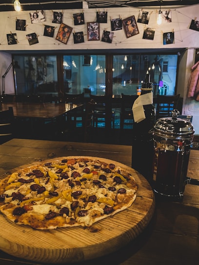 A thin-crust pizza topped with various ingredients including sausage, peppers, and cheese is placed on a wooden board. The setting is a dimly lit cafe, featuring hanging lightbulbs and an arrangement of photographs clipped to a string on the wall. A reflective French press and a dark bottle are situated next to the pizza.