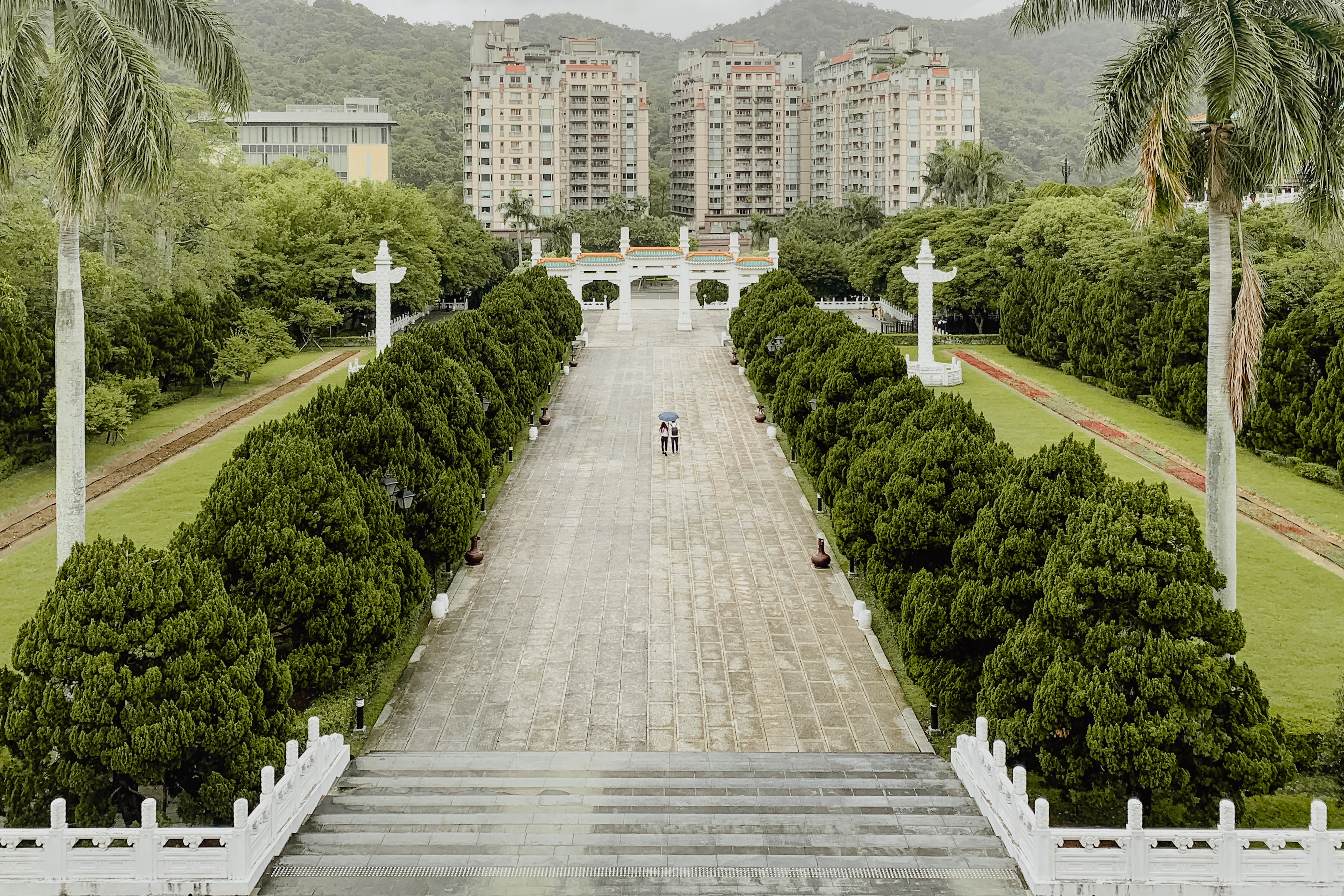 The approach to the National Palace Museum in Taipei seen from its steps on a grey damp day, a broad stone plaza flanked by clipped cypresses running down to the white ceremonial archway, two visitors sharing an umbrella at the centre and green hills behind.
