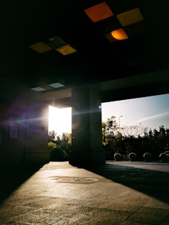Sunlight streams through an open doorway, casting long shadows on the textured floor. Trees and spherical bollards are visible outside, backlit by the sun. The ceiling above is adorned with various colored tiles, adding a geometric pattern to the otherwise dark interior.