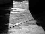 A black and white photograph features geometric shadows casting intricate patterns on a textured pavement. Strong contrasts between light and dark create a dramatic and abstract visual.