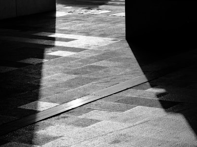 A black and white photograph features geometric shadows casting intricate patterns on a textured pavement. Strong contrasts between light and dark create a dramatic and abstract visual.