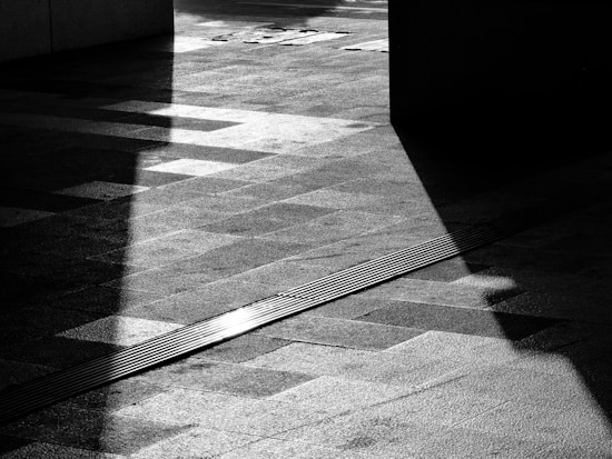 A black and white photograph features geometric shadows casting intricate patterns on a textured pavement. Strong contrasts between light and dark create a dramatic and abstract visual.