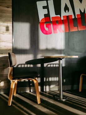 A wooden chair with a black seat cushion is positioned next to a small round table with a metal pedestal base. They're placed against a wall with the words 'FLAME GRILLED' displayed prominently, partly in red and white, partially out of view. The wall exhibits textured wooden and dark panels. Sunlight is casting strong shadows across the floor and the furniture, creating an interplay of light and darkness.
