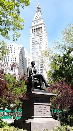 A statue of a seated figure is prominently displayed in a park surrounded by greenery. Behind the statue, a tall, white clock tower rises against a clear blue sky. The base of the statue includes inscriptions detailing the achievements of the person it commemorates.