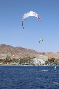 Two kite surfers glide across a deep blue sea with colorful kites soaring high in the clear sky. A picturesque beachfront town with white buildings, lush palm trees, and a mountainous backdrop is visible in the distance.