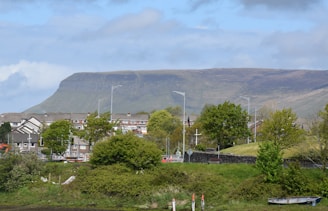 A residential area is situated in front of a wide and flat-topped mountain. The foreground features various green trees and shrubs, while street lamps line the street along the row of buildings. The sky is partly cloudy, creating a mix of light and shadow on the landscape.