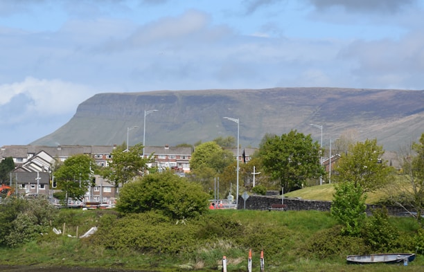 A residential area is situated in front of a wide and flat-topped mountain. The foreground features various green trees and shrubs, while street lamps line the street along the row of buildings. The sky is partly cloudy, creating a mix of light and shadow on the landscape.