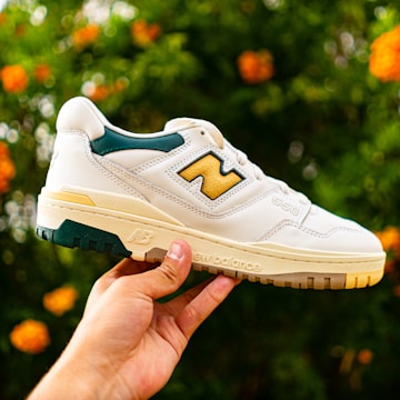 A hand is holding up a white sneaker with yellow and dark green accents against a blurred background of green foliage and orange flowers. The shoe features a prominent brand logo and text on the side.
