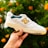 A hand is holding up a white sneaker with yellow and dark green accents against a blurred background of green foliage and orange flowers. The shoe features a prominent brand logo and text on the side.