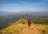A group of people are hiking along a narrow mountain ridge with lush green valleys stretching into the distance under a clear blue sky.