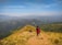 A group of people are hiking along a narrow mountain ridge with lush green valleys stretching into the distance under a clear blue sky.