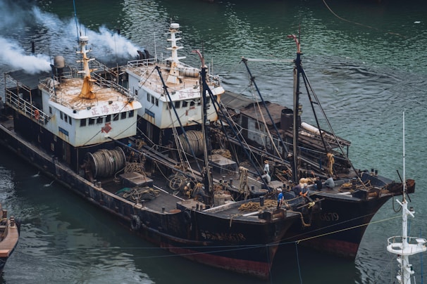 Two fishing vessels are docked side by side in a body of water, equipped with various fishing equipment and machinery on their decks. There is smoke emitting from one of the ships. Several crew members can be seen working on the decks of the boats. The water surrounding the boats has a greenish hue.