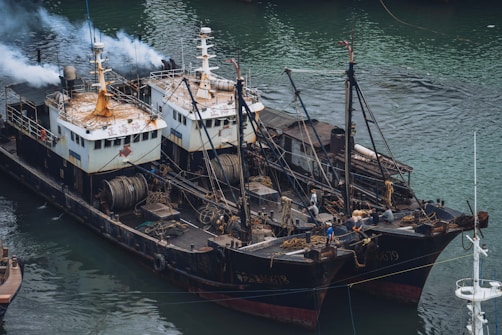 Two fishing vessels are docked side by side in a body of water, equipped with various fishing equipment and machinery on their decks. There is smoke emitting from one of the ships. Several crew members can be seen working on the decks of the boats. The water surrounding the boats has a greenish hue.