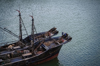 A large fishing boat floats on a calm body of water. The vessel is equipped with various fishing gear, ropes, and nets neatly arranged on deck. Two individuals are standing at the bow, appearing to be engaged in some activity related to the boat's operation.