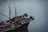 A large fishing boat floats on a calm body of water. The vessel is equipped with various fishing gear, ropes, and nets neatly arranged on deck. Two individuals are standing at the bow, appearing to be engaged in some activity related to the boat's operation.