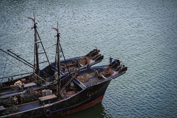 A large fishing boat floats on a calm body of water. The vessel is equipped with various fishing gear, ropes, and nets neatly arranged on deck. Two individuals are standing at the bow, appearing to be engaged in some activity related to the boat's operation.