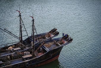 A large fishing boat floats on a calm body of water. The vessel is equipped with various fishing gear, ropes, and nets neatly arranged on deck. Two individuals are standing at the bow, appearing to be engaged in some activity related to the boat's operation.