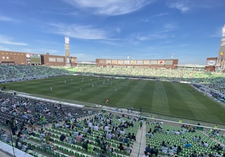 A large football stadium with a green field, partially filled with spectators seated in green and white seats. Players are on the field, and various advertisements are displayed around the stadium. The sky is clear with few clouds.