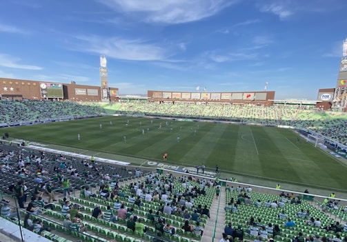 A large football stadium with a green field, partially filled with spectators seated in green and white seats. Players are on the field, and various advertisements are displayed around the stadium. The sky is clear with few clouds.