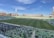 A large football stadium with a green field, partially filled with spectators seated in green and white seats. Players are on the field, and various advertisements are displayed around the stadium. The sky is clear with few clouds.