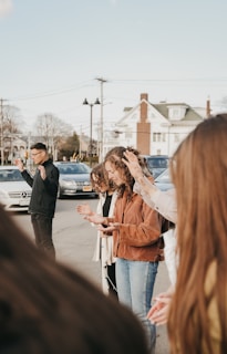 A group of people standing outdoors in a circle, actively engaged in an activity that involves raising their hands. Some individuals appear to be wearing casual winter clothing, and there are cars and a house in the background, with power lines overhead.