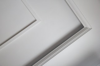 A close-up view of an interior ceiling corner with decorative molding. The light gray color of the wall contrasts slightly with the white trim, creating a subtle geometric pattern.